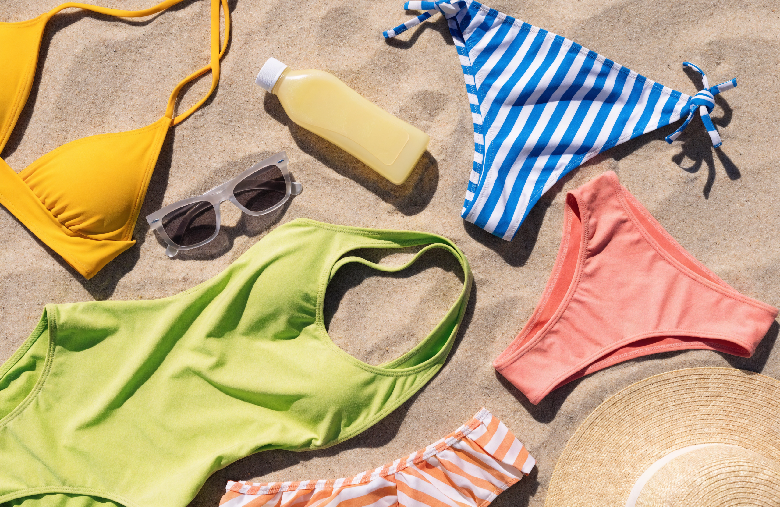 Illustrative flatlay of swimwear, sunglasses, sunscreen and a sun hat on sand